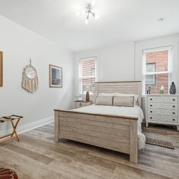 Bedroom with white walls, a wooden bed frame, two windows with blinds, a white dresser, and minimal decor.