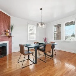 Bright dining room with white walls, hardwood floors, three large windows, a brick fireplace on the left, a modern chandelier, and a rectangular table with four chairs, decorated with a flower arrangement.