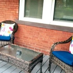 Outdoor patio with wicker furniture, a glass-top table, two chairs with cushions, and a brick wall with a window.