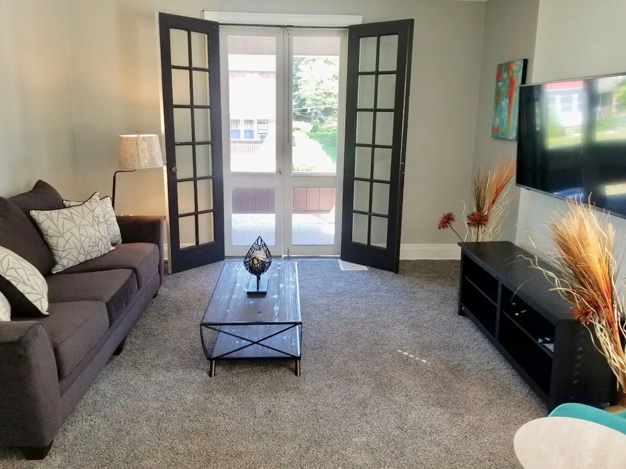 Living room with a brown sofa, decorative pillows, a lamp, a coffee table, a TV on a black console, and decorative plants, with French doors opening to a porch with greenery outside.