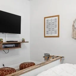 A bedroom corner with a wall-mounted TV, a floating wooden shelf, a framed chart, hanging decor, and a bed with a white headboard and bedding.