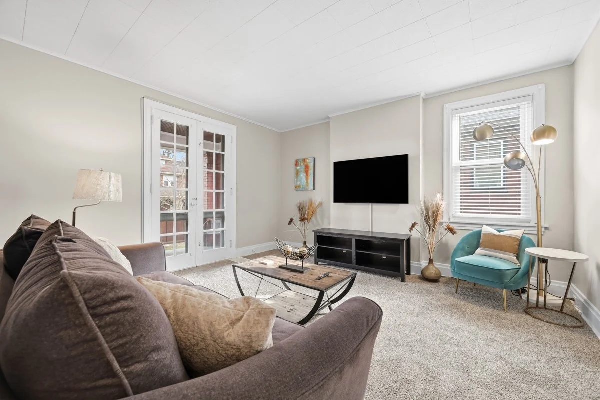 Living room with a brown sofa, beige pillows, a coffee table, a TV on a stand, and a teal armchair by the window with a small white side table, gold lamp, and decorative vases with dried plants.