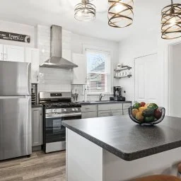 Modern kitchen with stainless steel appliances, a center island with a fruit bowl, white cabinets, a window with blinds, and pendant lights.