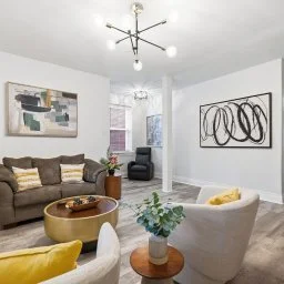 Modern living room with a brown sofa, white armchair, abstract artwork on the walls, and a gold coffee table. Bright space with a ceiling light fixture and large windows.