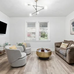 Living room with light gray armchairs, a dark brown sofa with pillows, a round coffee table, two windows with blinds, a TV mounted on the wall, and a modern ceiling light fixture.