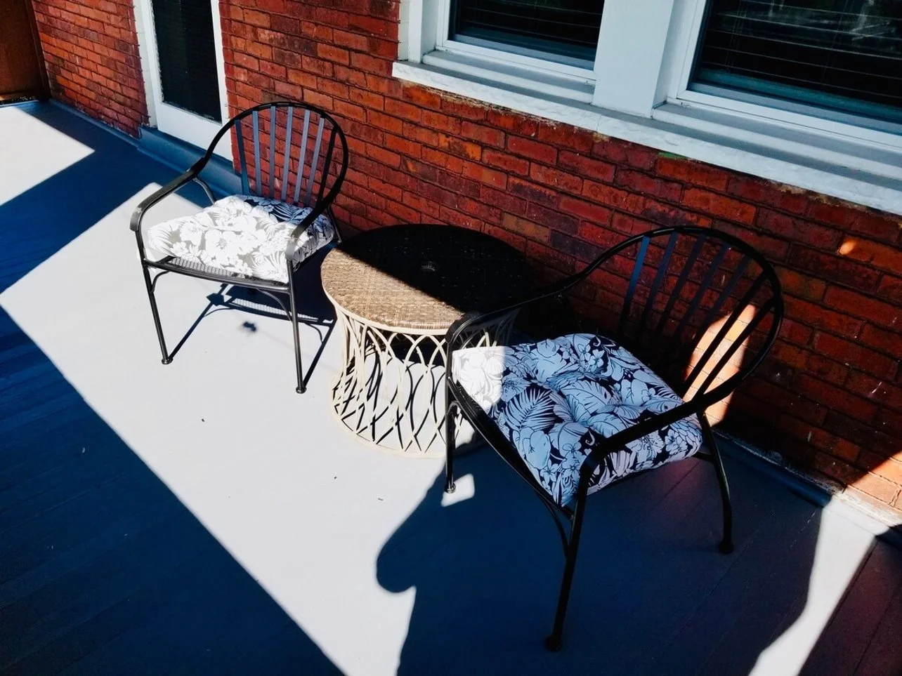 Two black outdoor chairs with patterned cushions on a porch with a brick wall and white window frames, separated by a round wicker side table, casting shadows in sunlight.