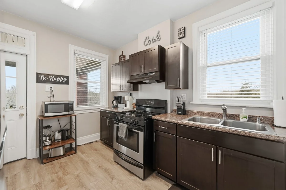 Kitchen with dark brown cabinets, a stainless steel stove and microwave, a granite countertop with a sink, a coffee maker, a paper towel roll, and decorative signs on the wall.