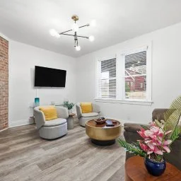 Modern living room with white walls, a brick accent wall, a flat-screen TV, yellow and gray armchairs, a round coffee table, a houseplant with pink flowers, and large windows with blinds.
