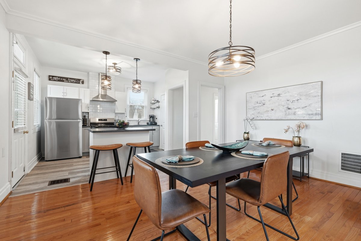 Dining area with a black table set with placemats and napkins, surrounded by brown leather chairs. A modern light fixture hangs above. The background features a kitchen with white cabinets, a stainless steel refrigerator, and a small breakfast bar wi