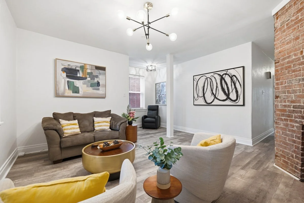 Living room with a brown sofa, white armchair with yellow pillow, black armchair, abstract artwork, modern chandelier, exposed brick wall, wooden flooring, and decorative plants.