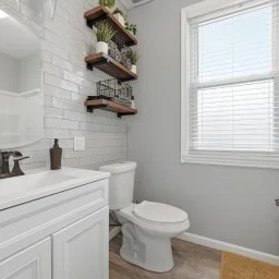 Bathroom with white vanity, toilet, wall-mounted shelves, a window with blinds, and a light-colored floor.