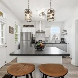 Kitchen with white cabinets, stainless steel appliances, and a central black countertop island with two wooden stools. Modern pendant lights hang above the island. There's a window with blinds and decorative shelves on the wall.