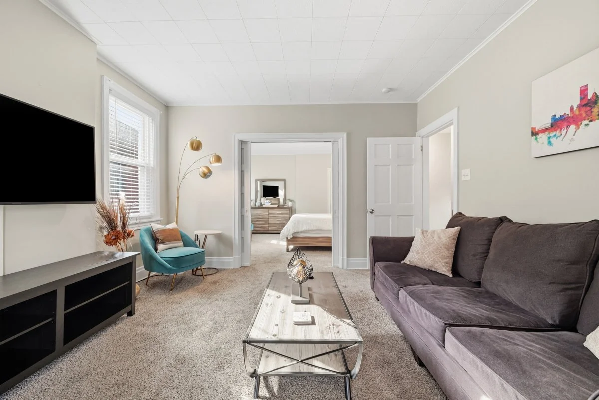 Living room with a dark purple sofa, a wooden coffee table, a teal armchair, a black TV on a stand, a floor lamp, and a painting on the wall. There is a doorway leading to a bedroom with a bed and a dresser.