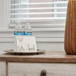 Two plastic water bottles on a tray, with a notepad and pencil, placed on a wooden surface near a window with blinds and curtains.
