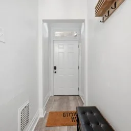 Entryway with a white front door, coat hooks on the wall, a black bench, and a orange welcome mat on wooden floor.