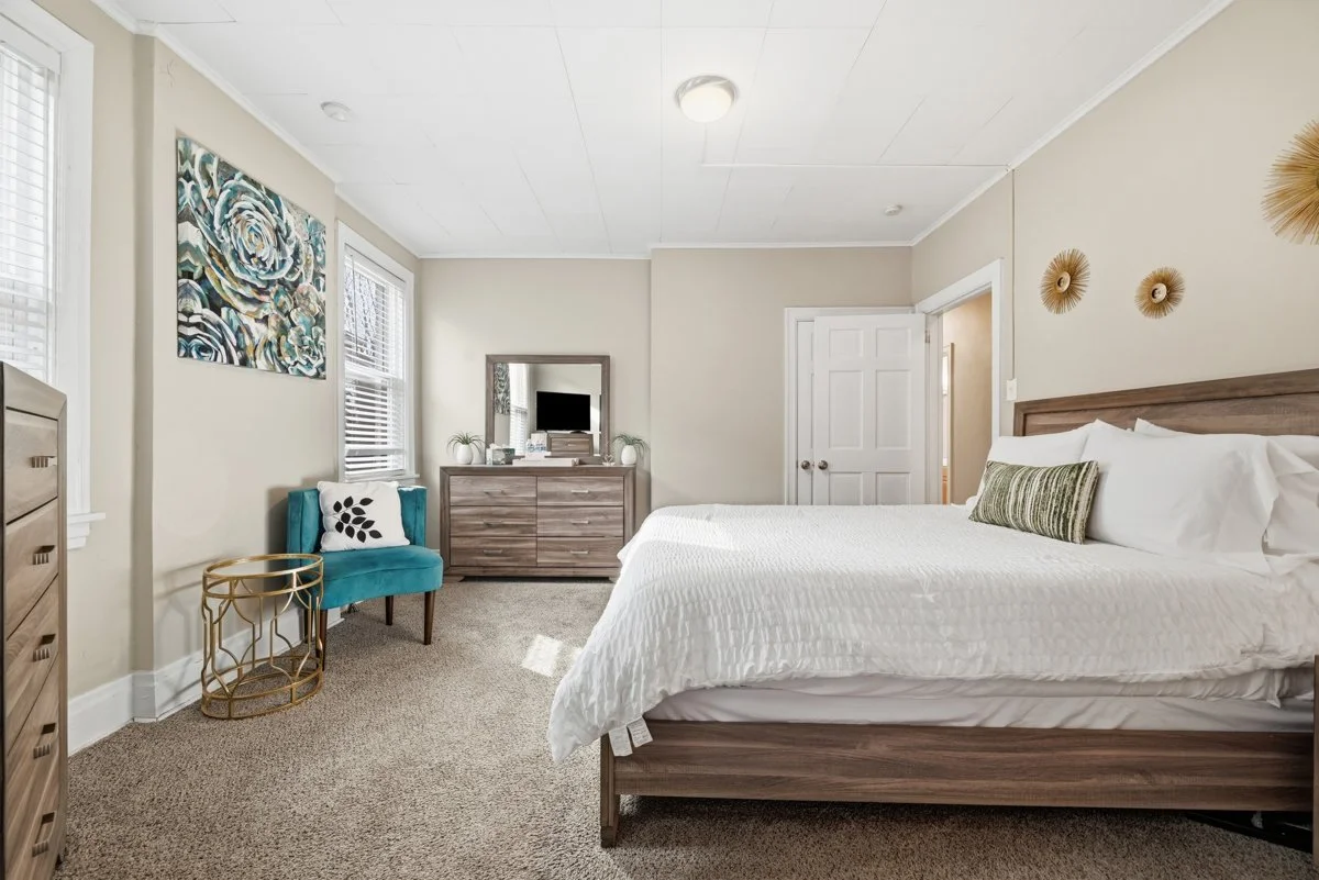 A bedroom with a large bed, adorned with white bedding and a green striped pillow. Opposite the bed, there's a wooden dresser with a mirror, decorated with small plants, books, and a TV. To the left, a blue armchair with a black and white pillow is n