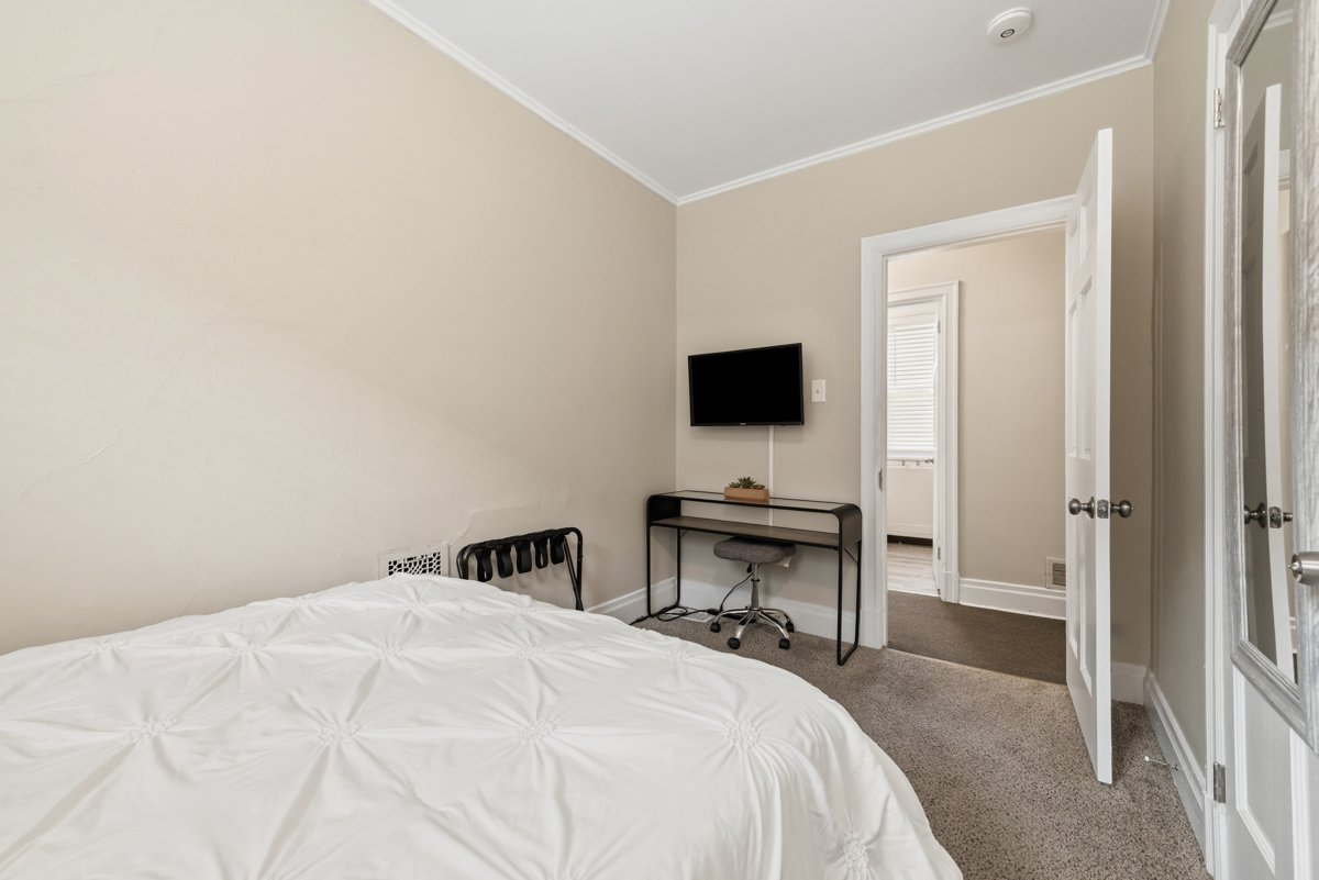 A bedroom with beige walls, a bed with white bedding, a small desk with a chair beneath a wall-mounted TV, and an open door leading to another room with a window.