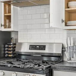 Kitchen with white tiled backsplash, stainless steel stove and oven, open wooden shelves, and countertop with dishes, knives, and a spice rack.