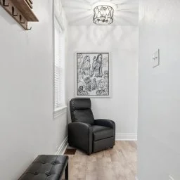 Small white room with a black leather armchair, a textured black ottoman, a window with blinds, a framed black-and-white artwork, and a ceiling light fixture.