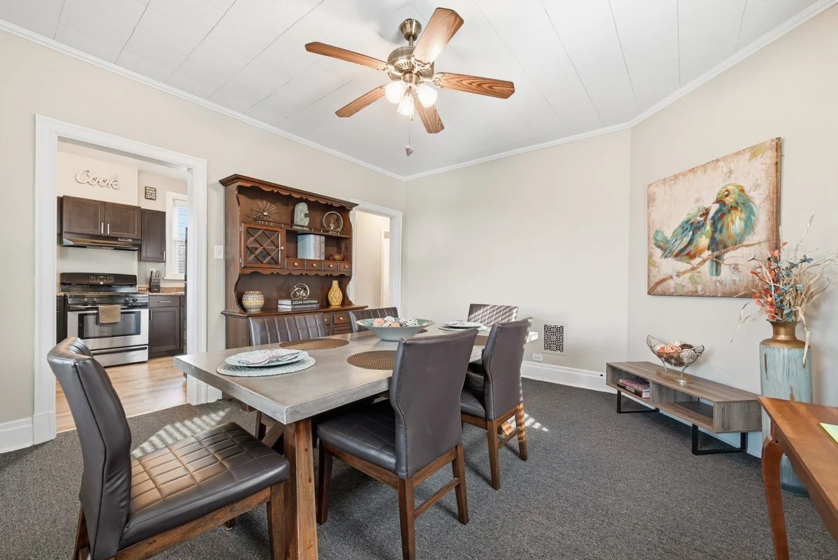 A dining room with a wooden table and six leather chairs, a wooden hutch with decorative items, a colorful bird painting on the wall, a vase with dried flowers, a TV stand, and a ceiling fan with lights, with a doorway leading to a kitchen.