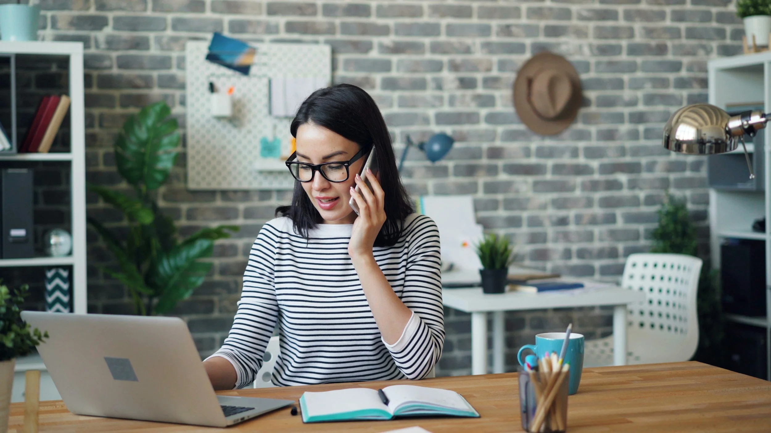 A woman with dark hair, glasses, and a striped shirt talking on the phone while working on a laptop at a wooden desk. The desk has an open notebook, a pen, a mug, and a container of pens. The background shows a brick wall, plants, and shelves with books and decorative items.