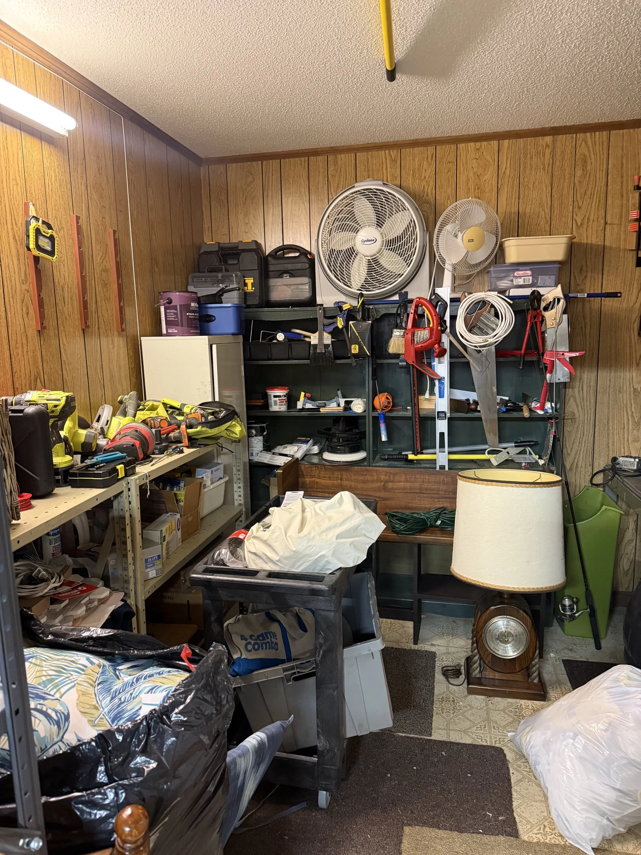 A cluttered  basement with wooden paneled walls and a mixture of tools, storage bins, and household items that was decluttered and then organized with various power tools on a workbench, and a pile of bags and boxes on the floor.