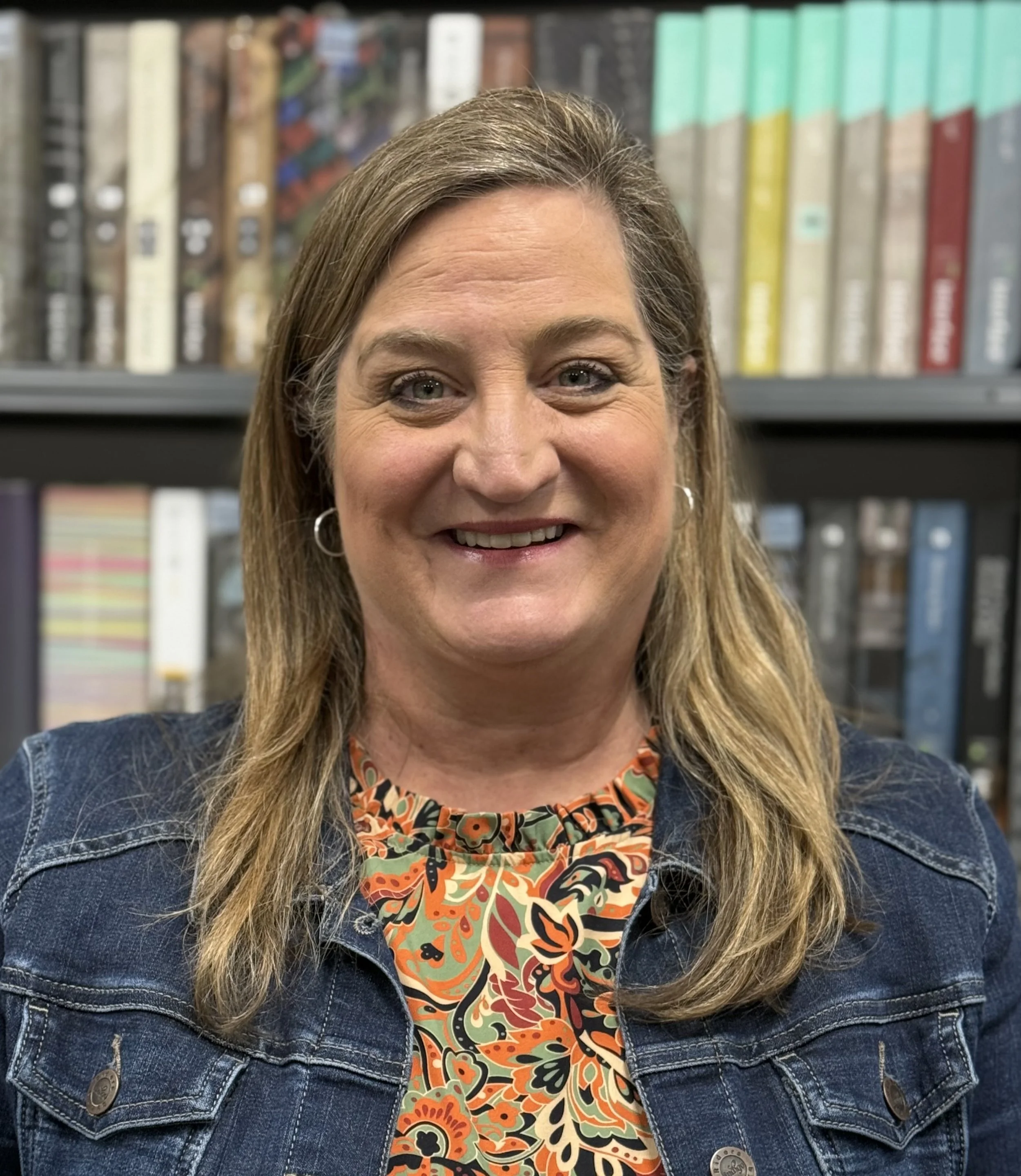 A woman with long blonde hair, wearing a colorful floral shirt and denim jacket, smiling in front of a bookshelf.