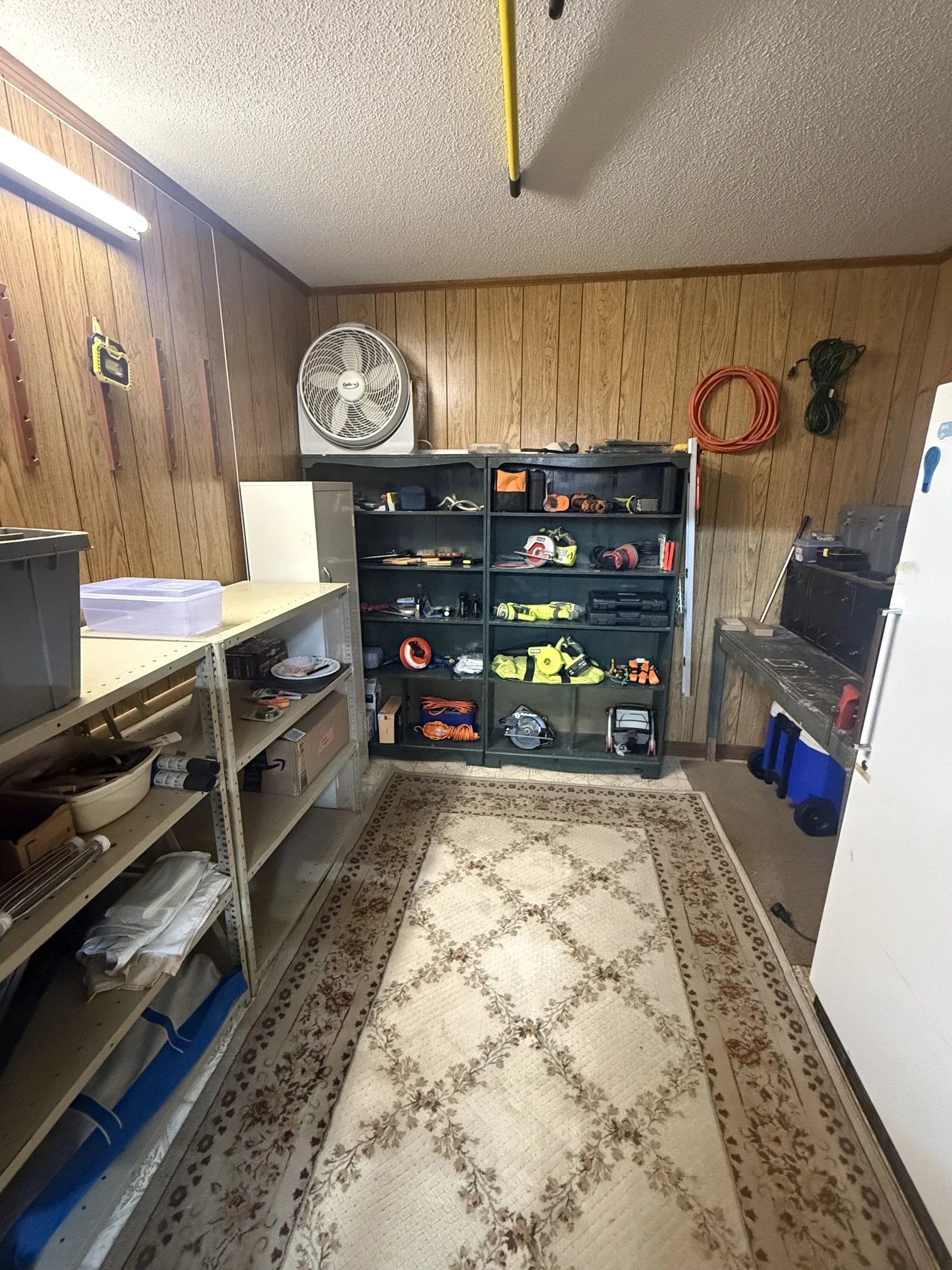 The image shows a garage or workshop with wood-paneled walls, a beige patterned rug on the floor, and various tools and equipment organized on shelves. There is a fan on the top shelf and a collection of power tools and supplies in a black shelving unit. A white countertop with miscellaneous items is on the left, and a white refrigerator or cabinet is on the right.