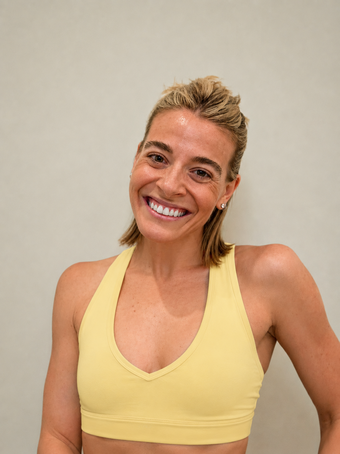 A woman with long light brown hair seated in front of a black wall with vertical stripes, with a neon orange yin-yang sign above her head. She is wearing a brown tank top and matching brown pants, smiling slightly, with her hands resting on her legs.