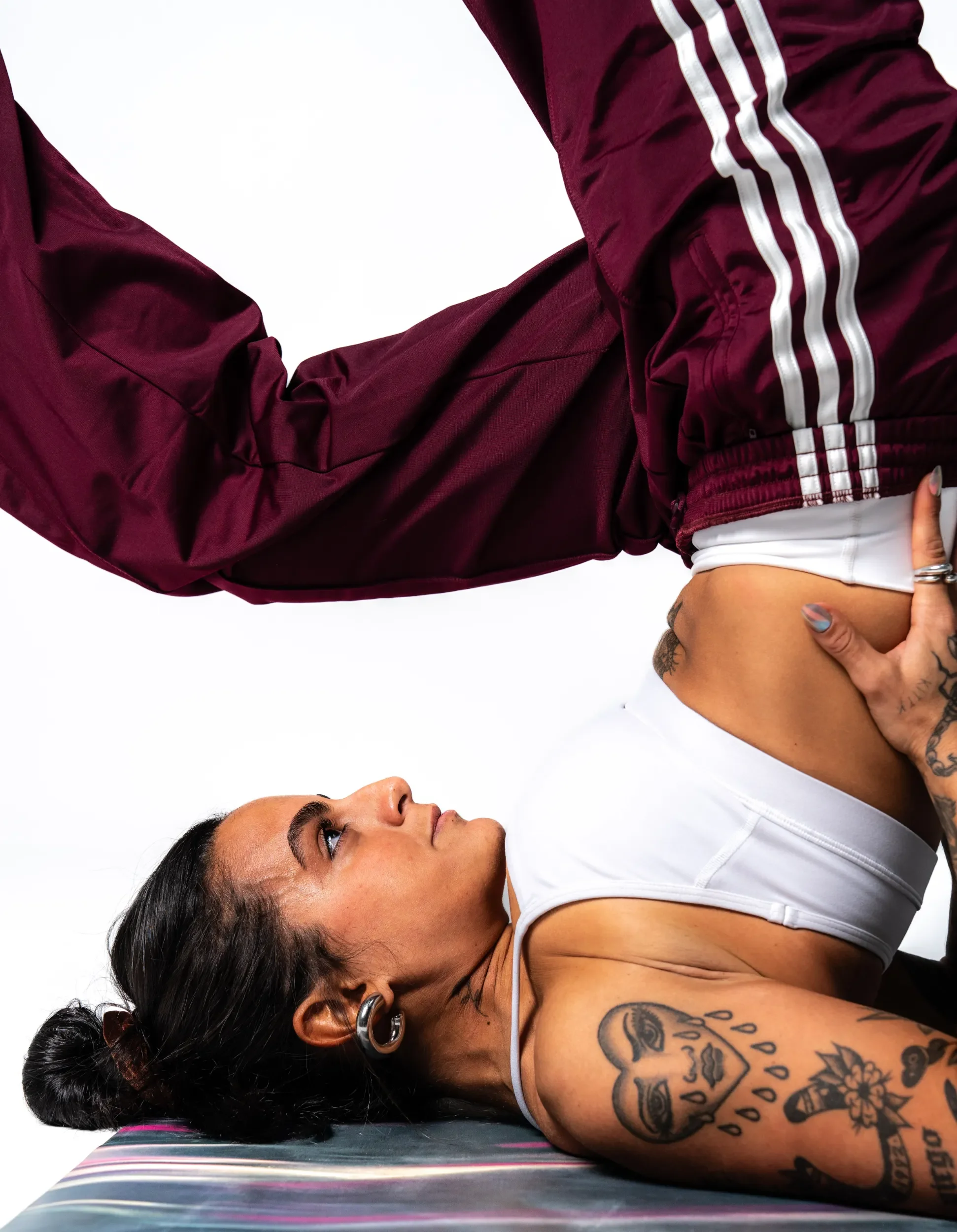 A woman with dark hair in a bun and tattoos on her arm lying on a metallic exam table, looking upward, wearing a white tank top.