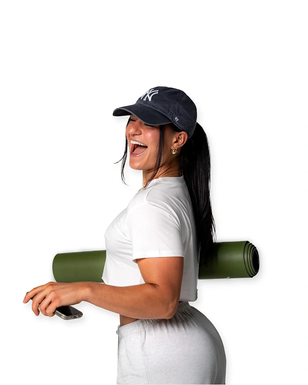 Woman in white shirt and gray pants with a New York Yankees cap, holding a smartphone in her hand and a green yoga mat on her shoulder, smiling and laughing.