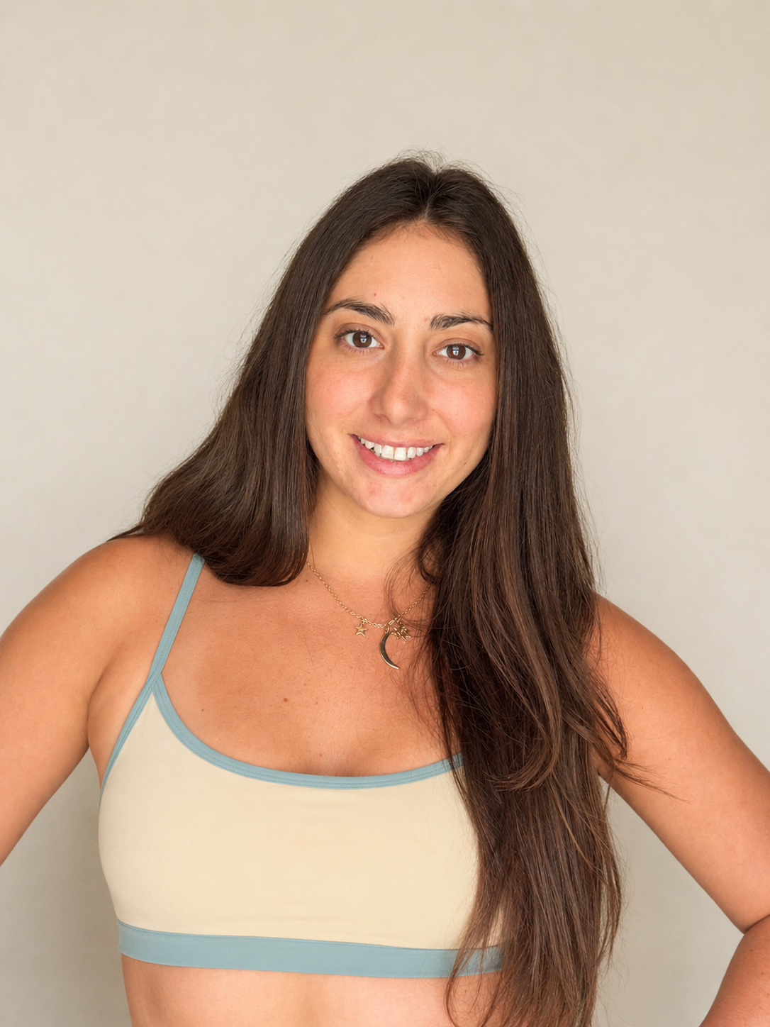 A woman with long, light brown hair, wearing a black bikini top and black shorts, standing in front of a white door, smiling with her hands on her hips.