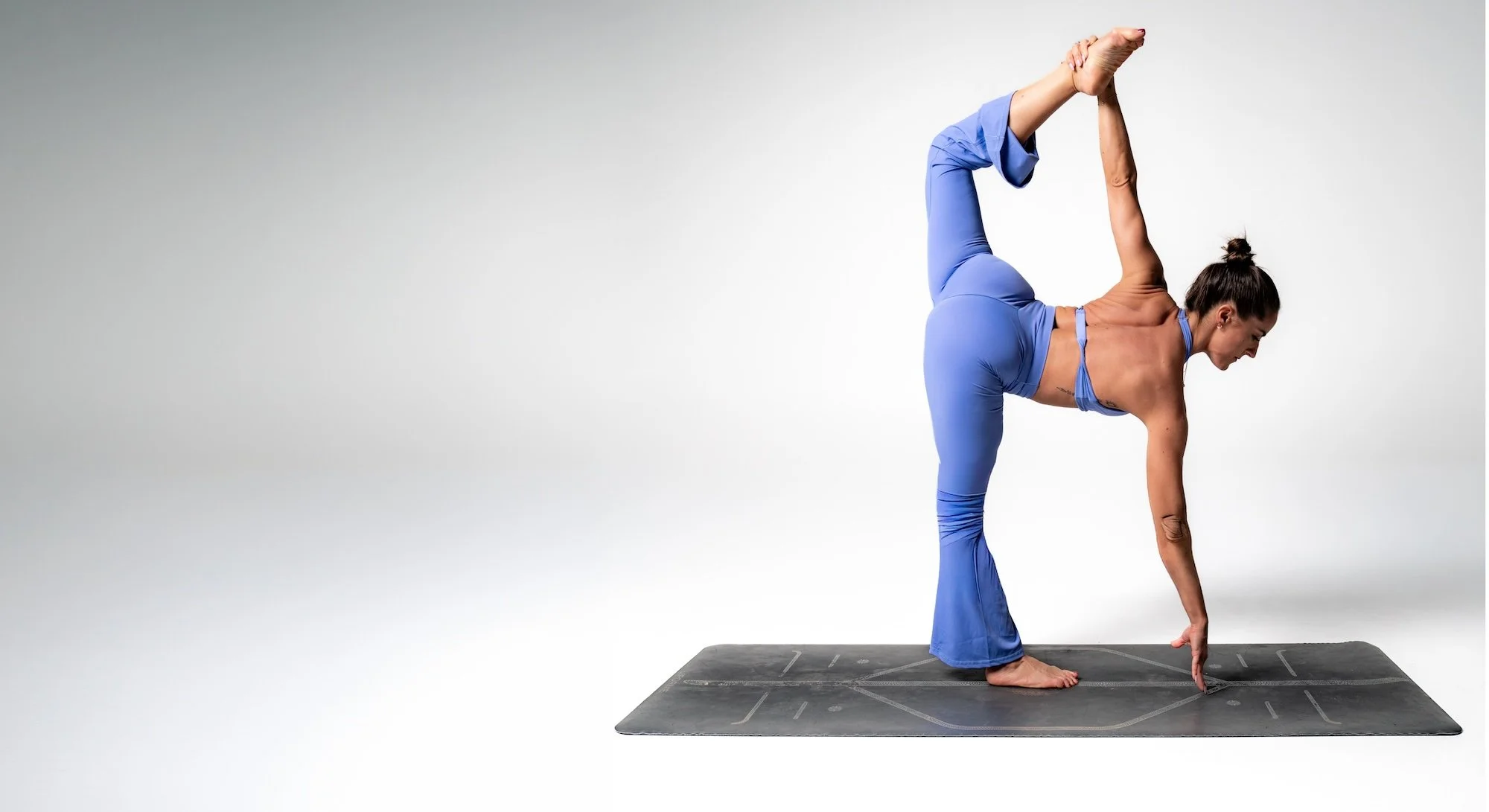 Woman in blue exercise clothes performing a yoga pose on a black mat against a plain light gray background