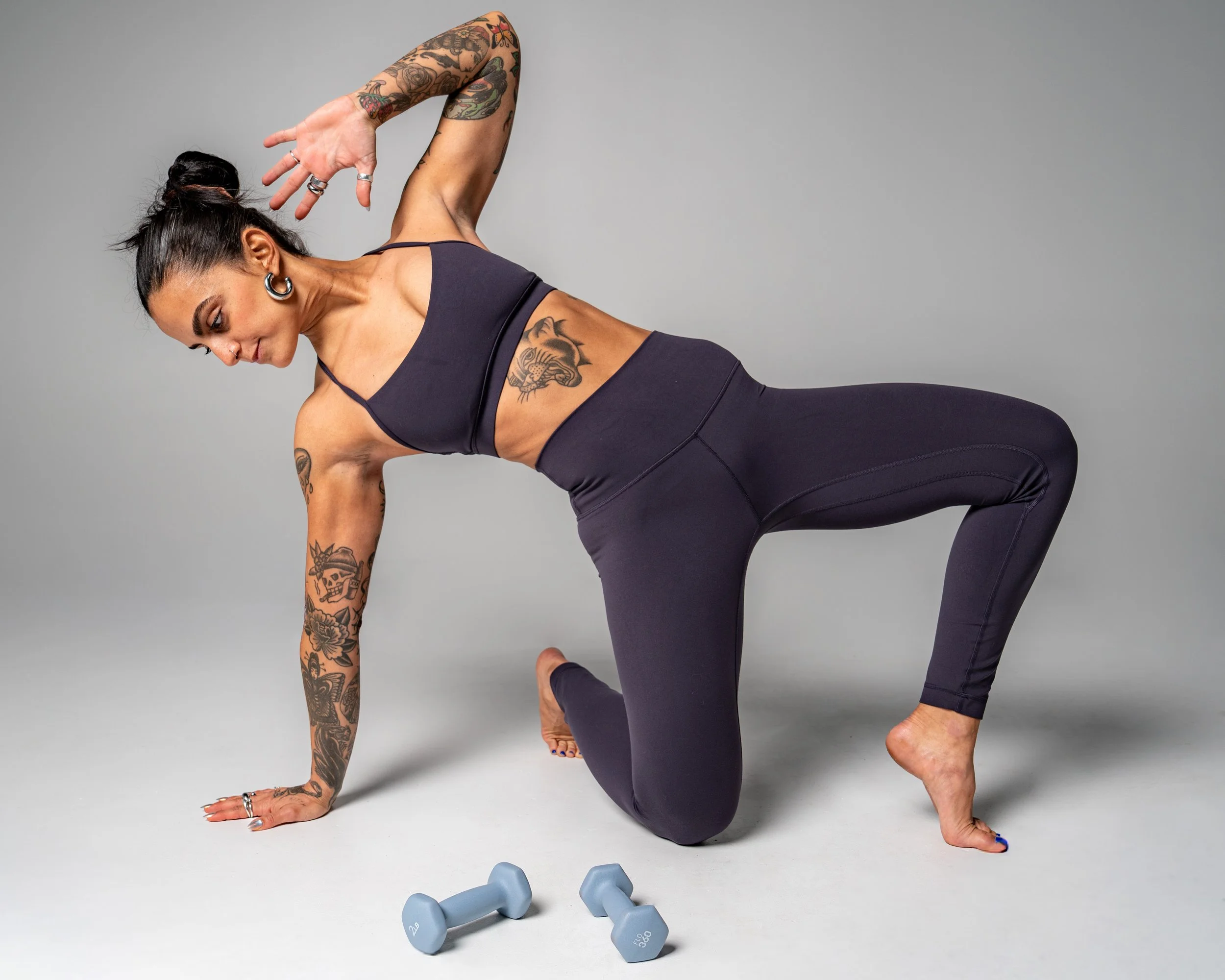 A woman with tattoos on her arms and torso, wearing black athletic wear, is in a yoga pose on a light gray background. She is balancing on her hands and feet with her back arched.