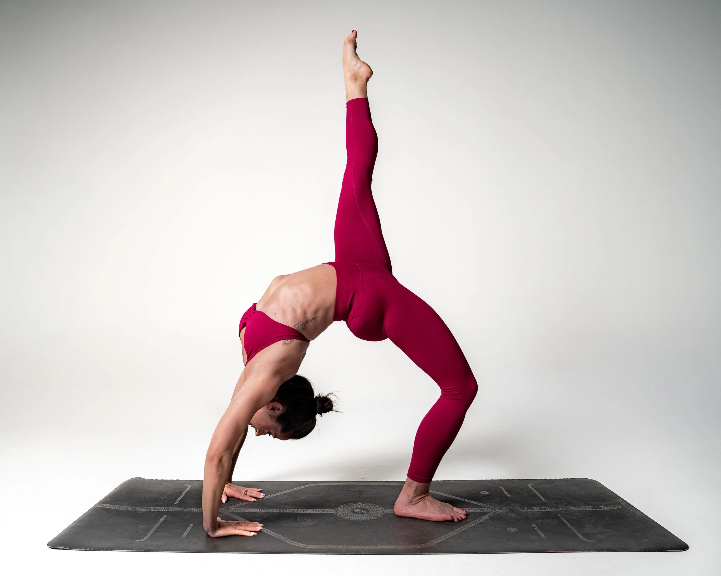 A woman in pink workout clothes performing a yoga pose called the wheel pose or upward bow on a black yoga mat in a studio with a plain white wall