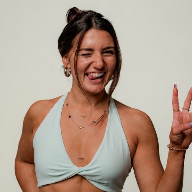 A young woman with brown hair styled in a messy bun, wearing jewelry and a light blue sports bra, sticking out her tongue and making a peace sign with her right hand, against a plain background.
