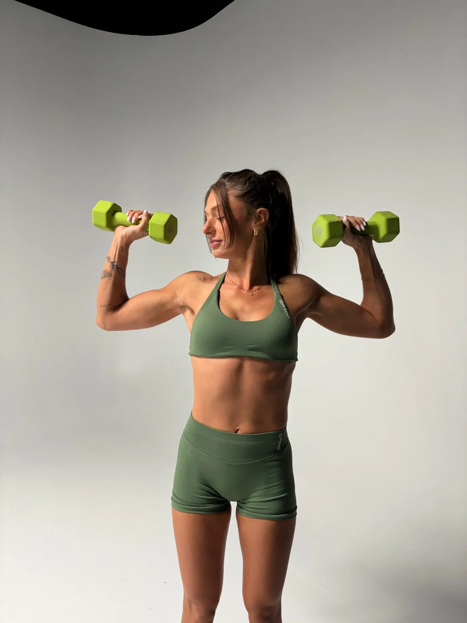 A woman in green workout attire lifting green dumbbells in a studio with a plain light gray background.