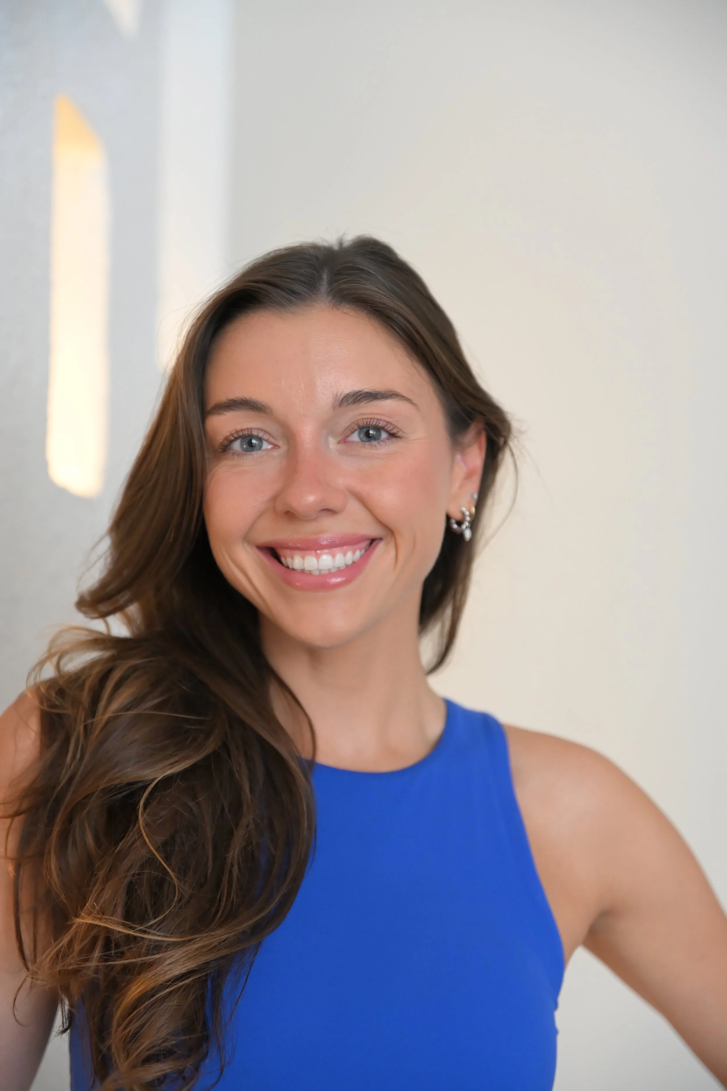 A woman with long, light brown hair, wearing a black bikini top and black shorts, standing in front of a white door, smiling with her hands on her hips.