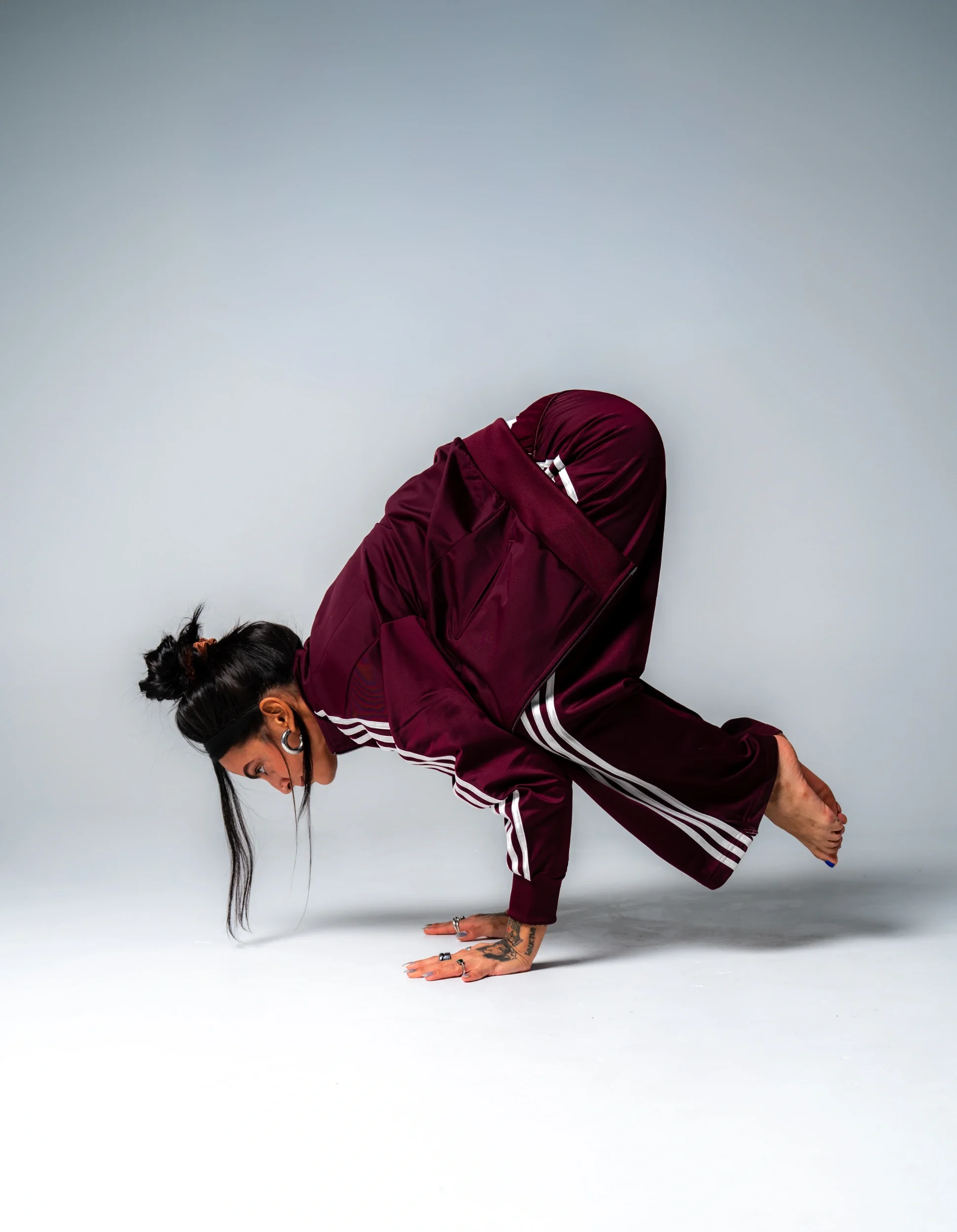 A woman in a maroon athletic jacket and pants is performing a yoga pose called the handstand split, balancing on her hands with her legs extended in opposite directions against a plain white background.