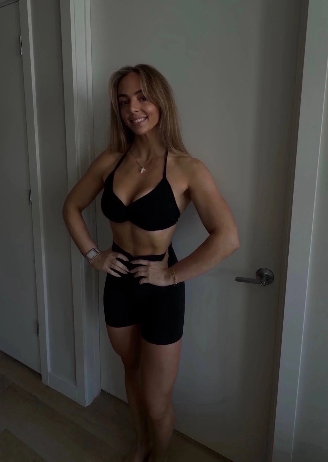 A woman with long, light brown hair, wearing a black bikini top and black shorts, standing in front of a white door, smiling with her hands on her hips.