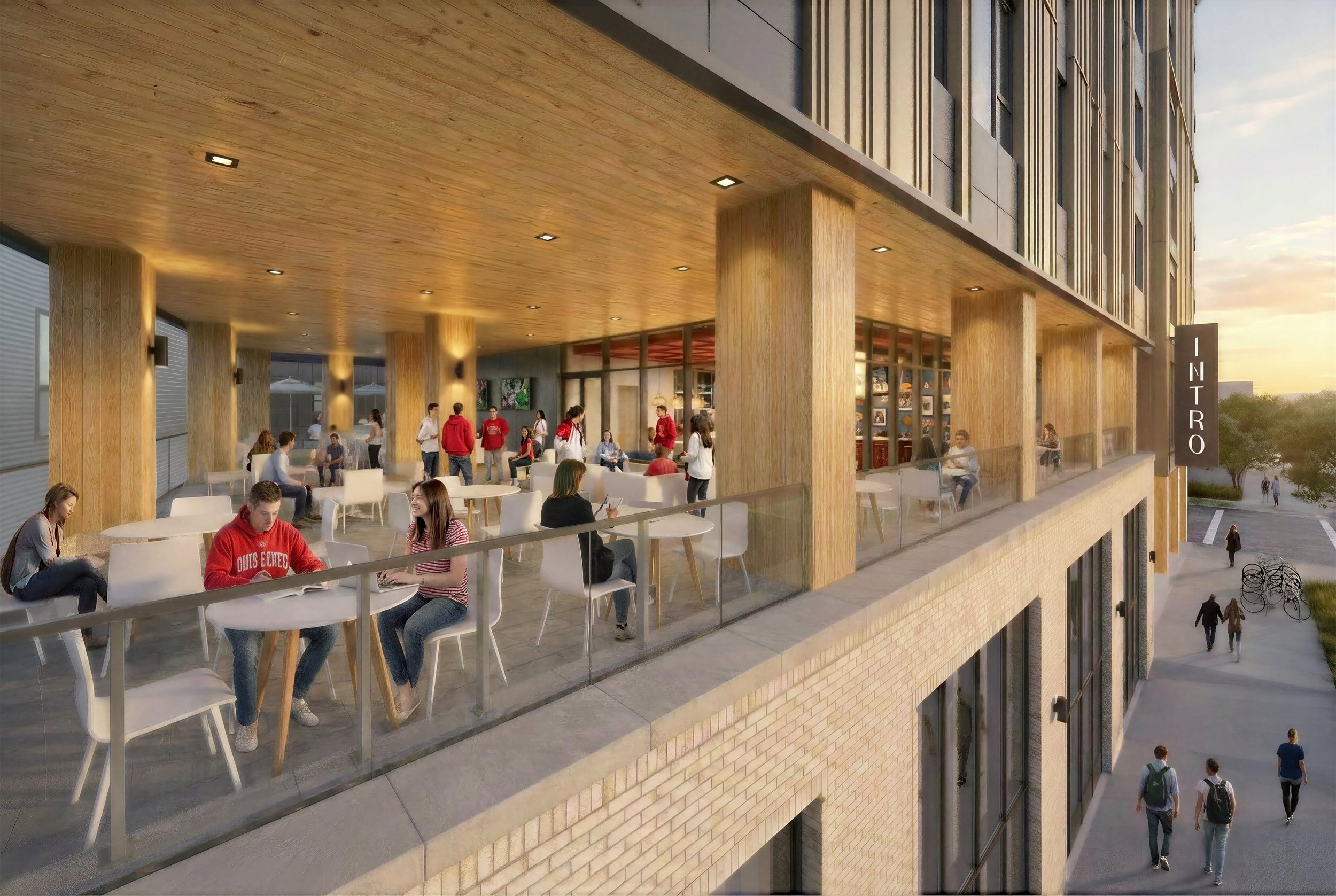 Exterior view of a modern building with a balcony where people are sitting and standing, some talking, at sunset. The building features wooden accents and large glass windows.