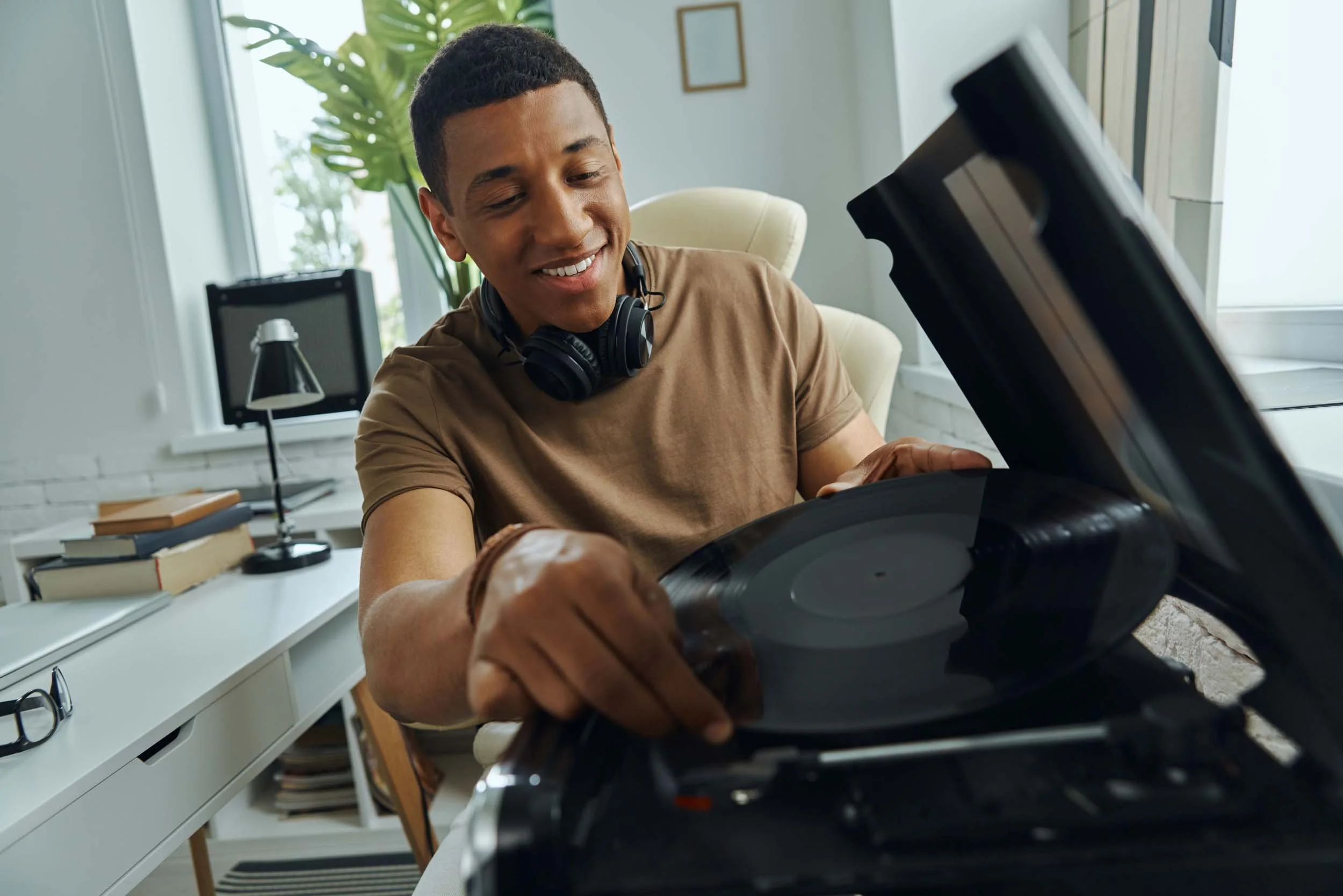 A smiling young man with short dark hair, wearing a brown t-shirt and black over-ear headphones around his neck, is opening the lid of a black record player on a white desk in a bright room with large windows, a green plant, and framed artwork in the background.