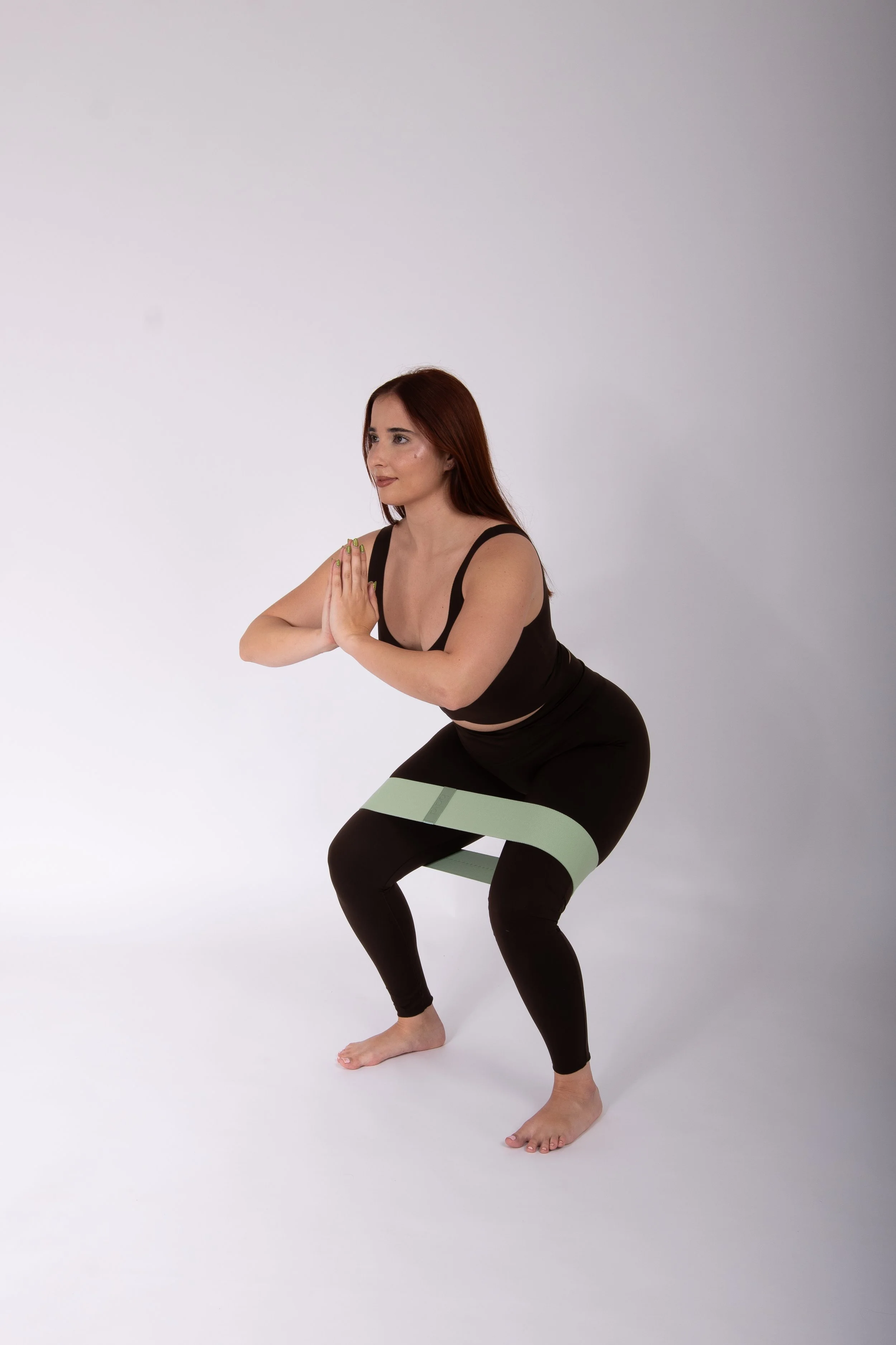 Mujer joven realizando ejercicio de yoga en un estudio con fondo gris claro, usando ropa deportiva negra, en posición de cuclillas con manos en oración.