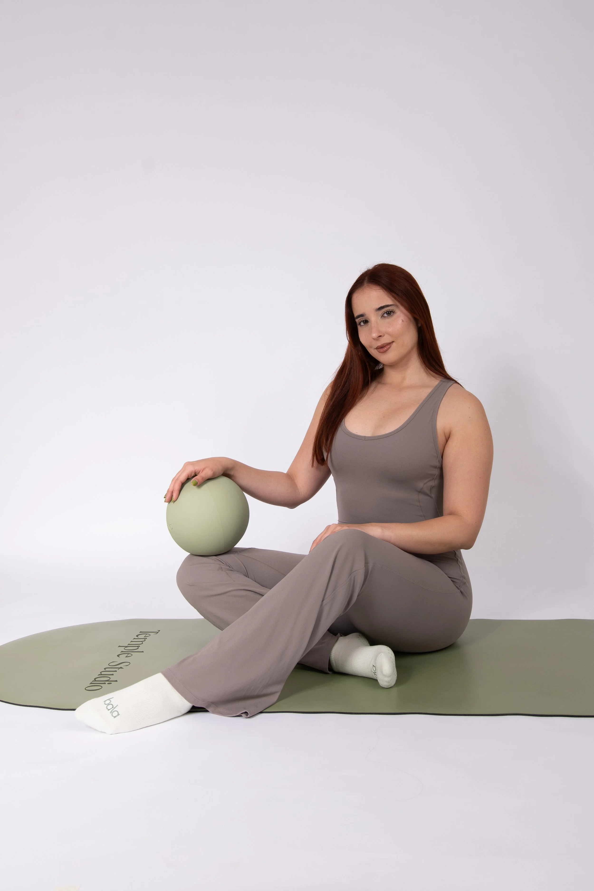 Mujer con cabello largo y rubio, vestida con ropa gris, sentada en un tapete de ejercicio, con una pelota en el regazo, en un entorno blanco, con expresión relajada y rostro sonriente.