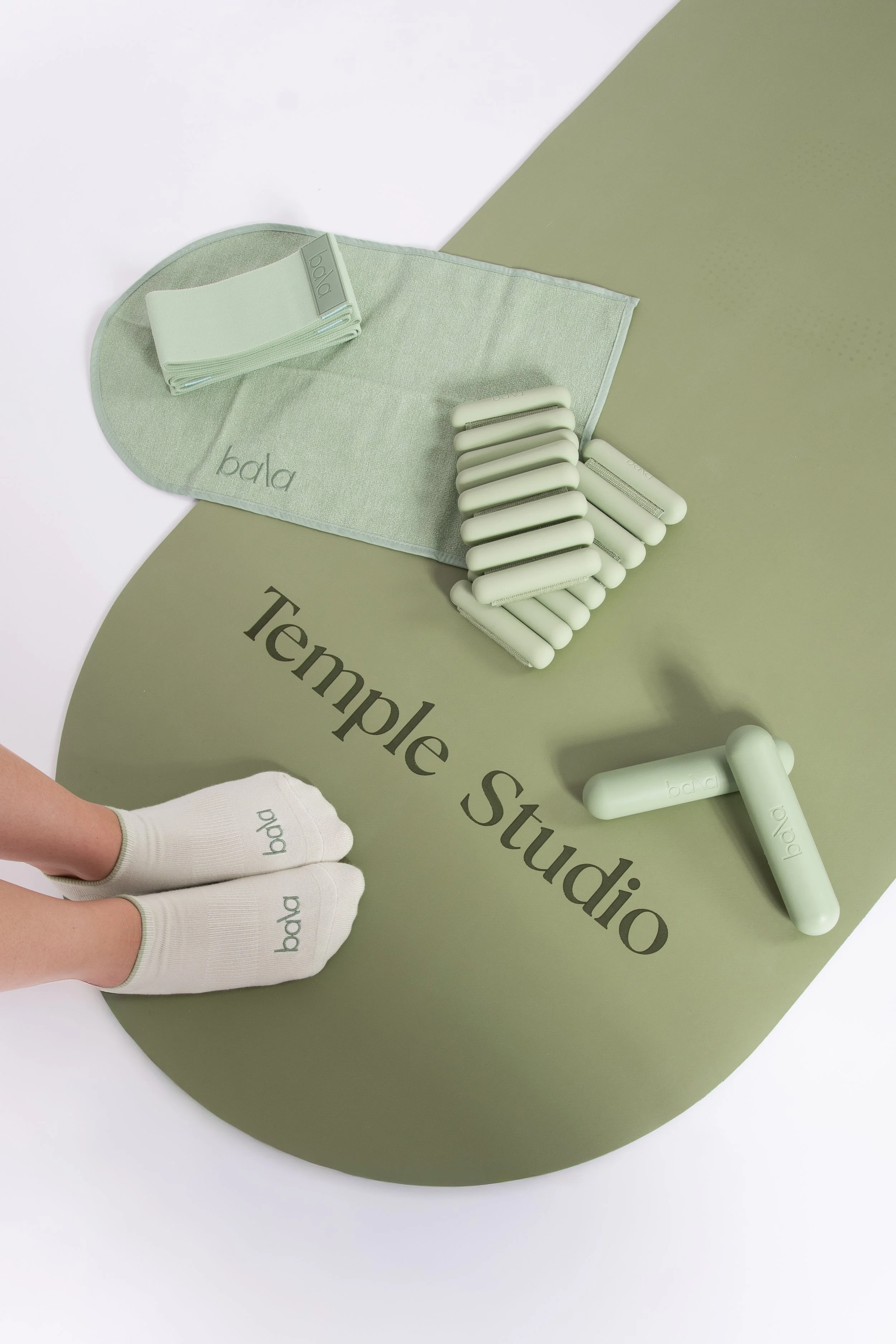 Manos con guantes blancos con la palabra 'bala' en la parte superior, sobre una mesa decorada en tonos verdes y blancos con objetos de higiene personal, incluyendo bloques de jabón y una toalla, y con palabras 'Temple Studio' en la mesa.