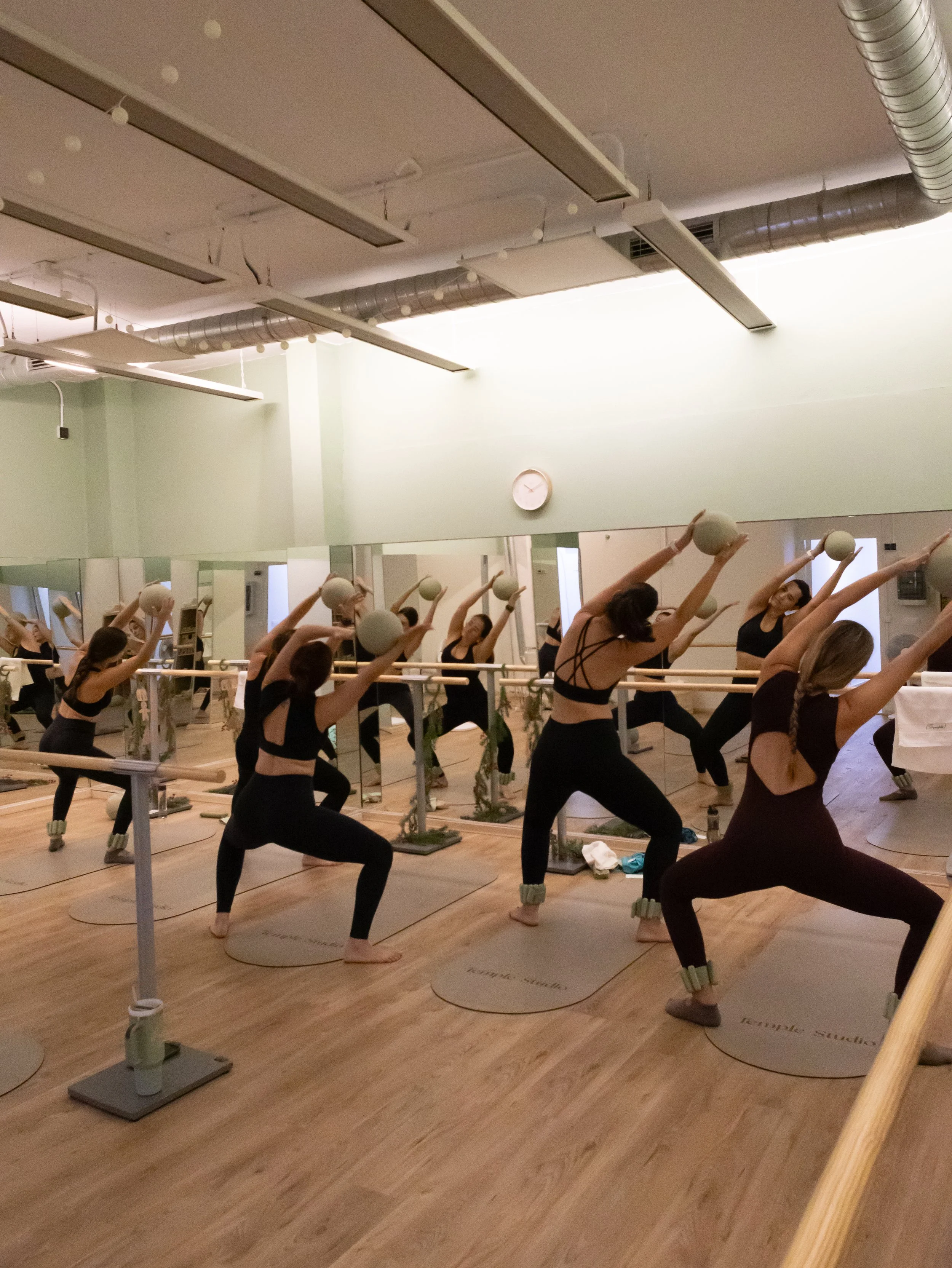 Grupo de personas haciendo ejercicio de yoga con bolas en un estudio de yoga con espejos y piso de madera.