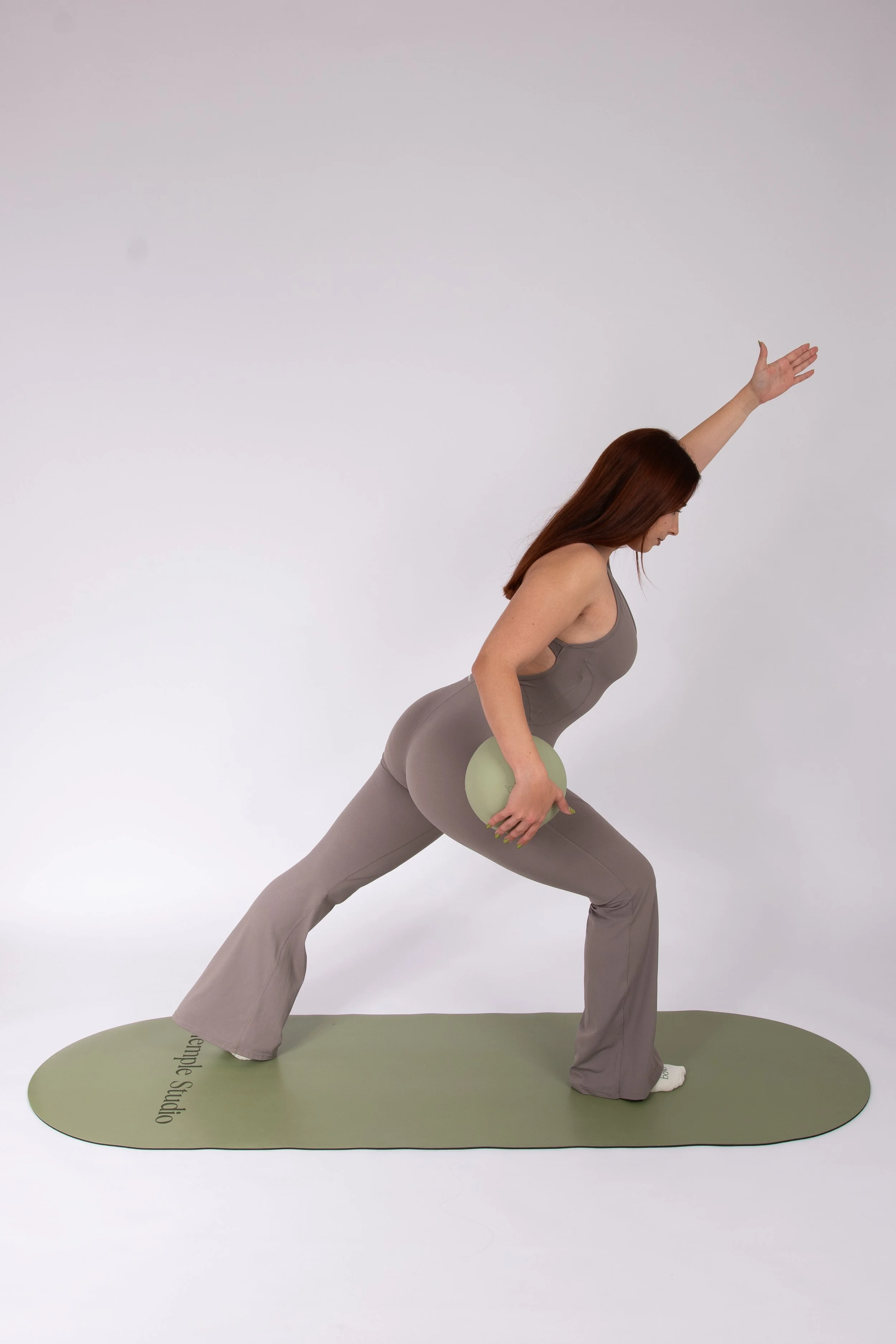 Mujer realizando yoga en una pose de estocada con peso, sobre esterilla verde, en un fondo gris.