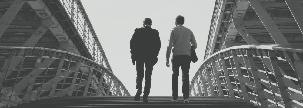 Two men walk up an outdoor staircase between modern buildings in black and white.