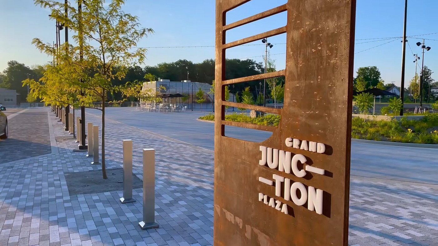 A sign reading "Grand Junction Plaza" next to a paved walkway with young trees and bollards, with an open plaza area and tables in the background, under a clear sky.