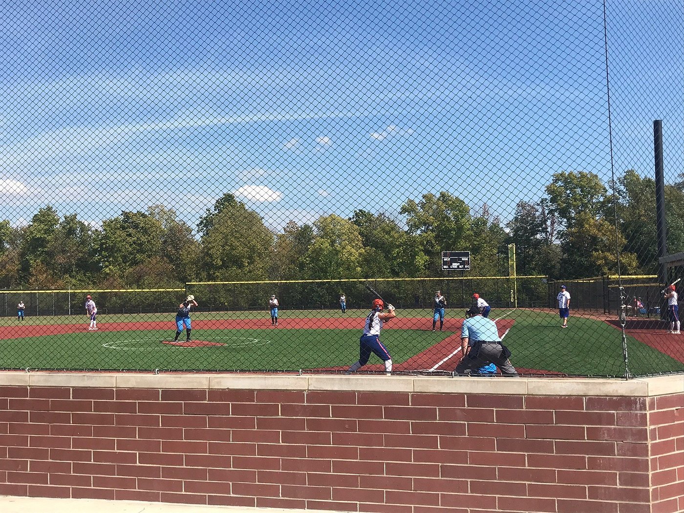 Grand Park Sports Campus baseball game with players in uniforms on the field, seen through a chain-link fence, with a blue sky and trees in the background.